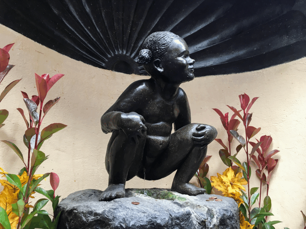 Jeanneke Pis, lille bronzestatue i Bruxelles, pige der tisser, pendant til Manneken Pis, moderne belgisk kunst, humoristisk seværdighed i Belgien, Jeanneke Pis i Bruxelles’ centrum, skjult seværdighed i en gyde, belgisk satire og kultur, sjove oplevelser i Bruxelles, symbol på ligestilling i kunst, quirky Brussels statue, Jeanneke Pis attraktion, must-see i Bruxelles, belgisk tradition i moderne form, unikke statuer i Belgien